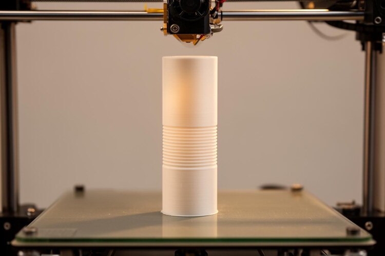 Высокий тонкостенный белый корпус печатается на столе 3D-принтера, видны слои и сопло над верхней кромкой